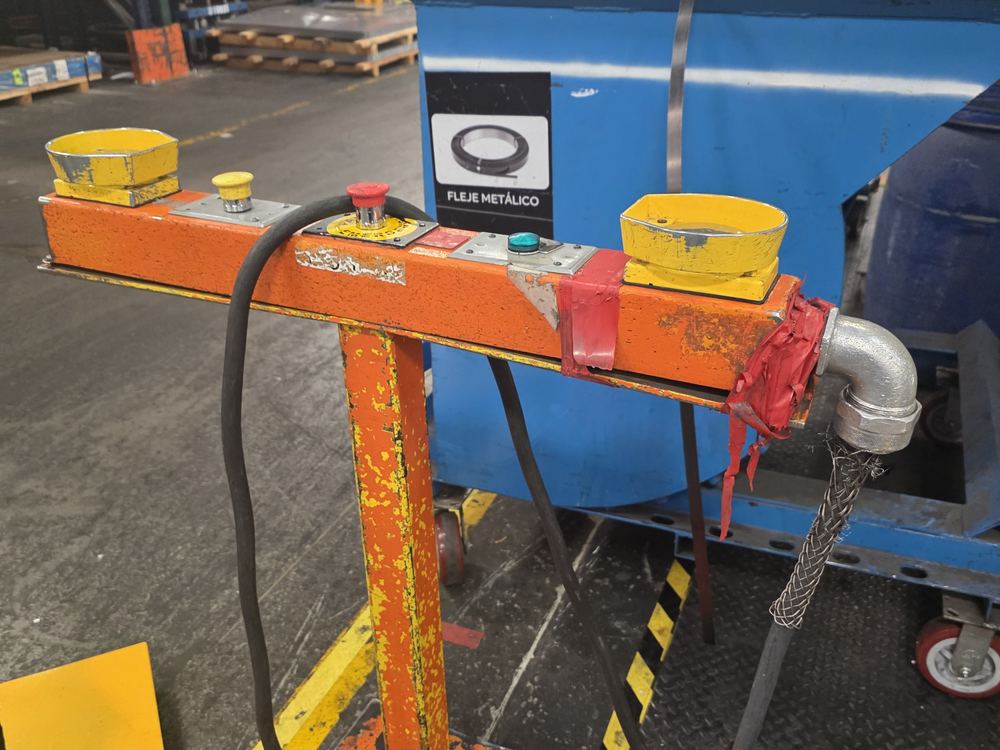 Estación de mando anterior: viga naranja oxidada sobre un poste con ruedas, con dos botones de palma, paro de emergencia y el cable tendido por el piso