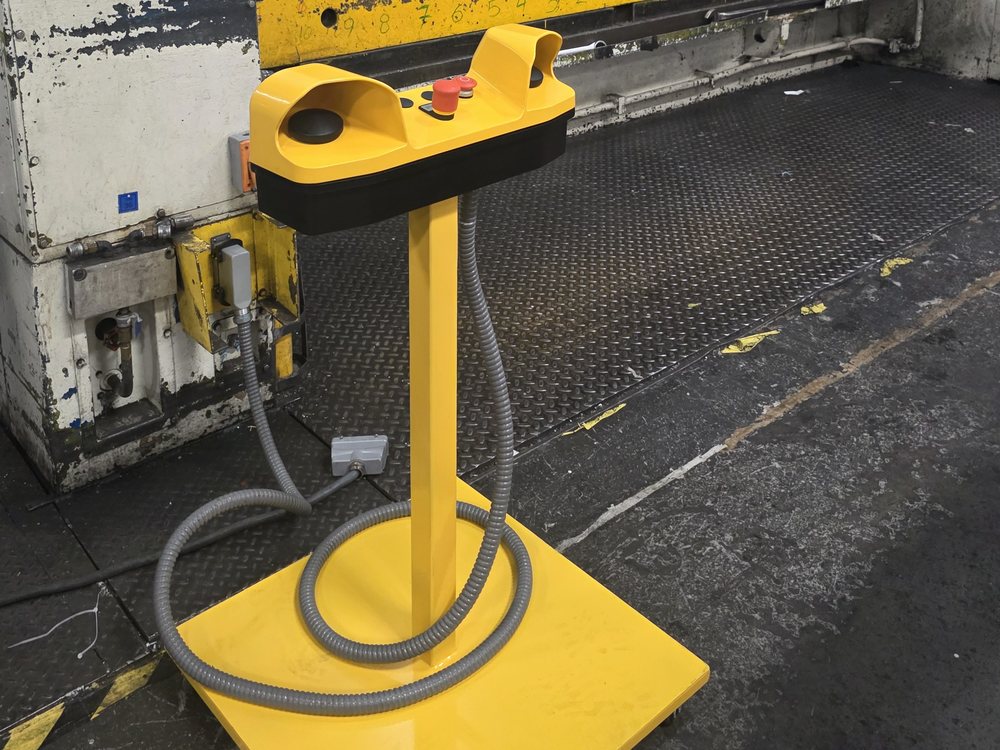 Estación de mando nueva: pedestal amarillo con cabezal de mando a dos manos y paro de emergencia, instalado frente a una prensa