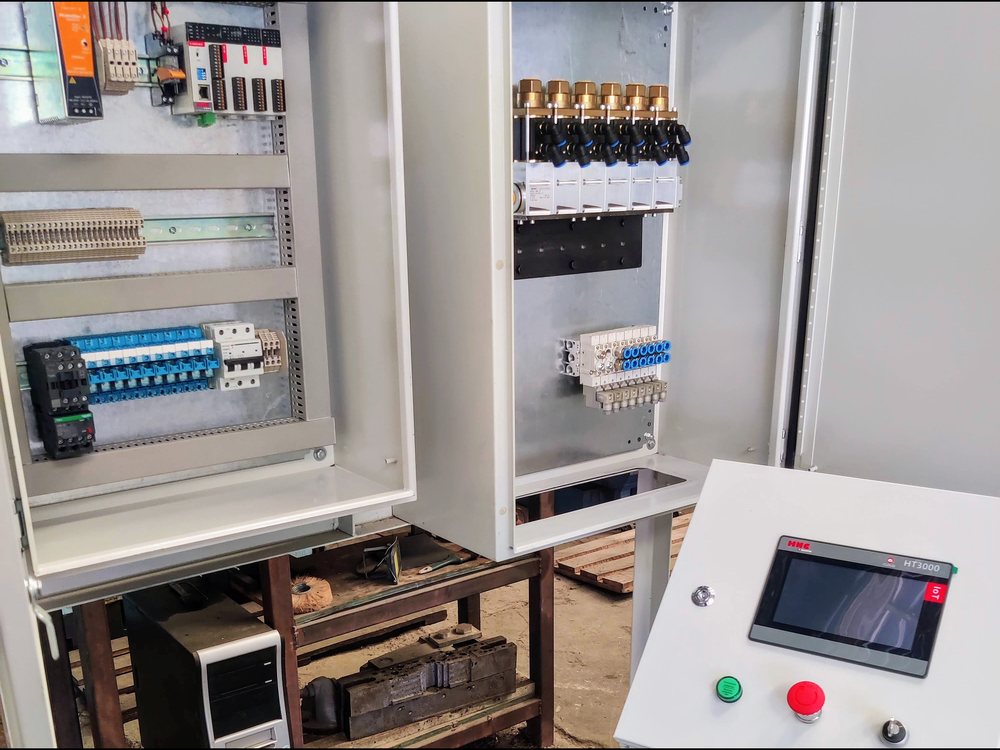 Tablero de control de la estación de prueba de estallido: dos gabinetes abiertos con PLC, protecciones y bornes, manifold de válvulas neumáticas y pedestal con pantalla HMI